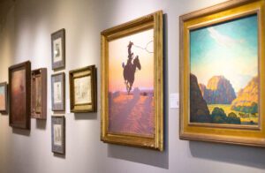 A wall of photos in a museum. The picture being focused on shows a cowboy throwing a lasso while riding a horse in mid action.