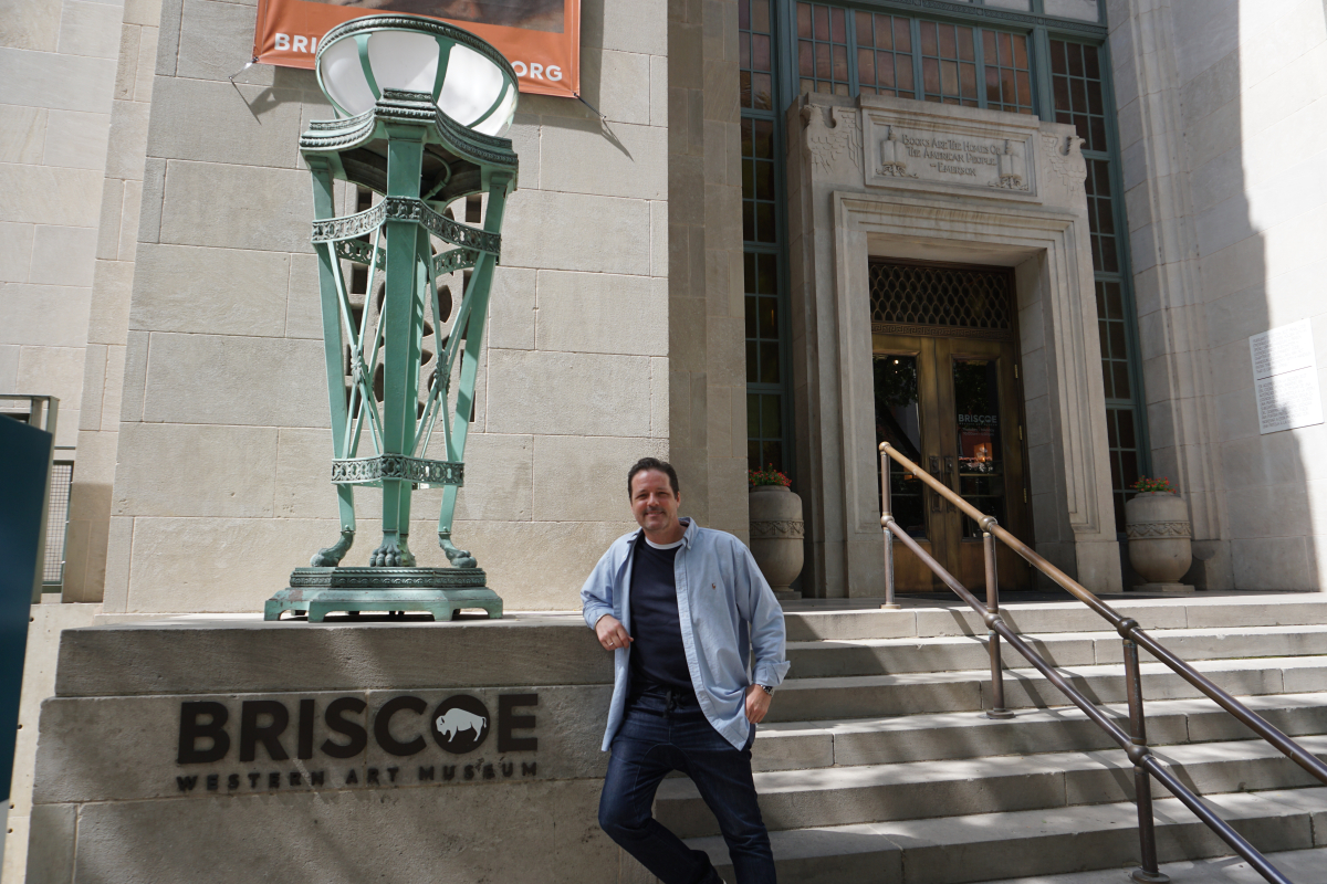 An individual standing in front of the Briscoe Western Art Museum. The person is next to a large, ornate lamp post, and the museum entrance features steps leading up to double doors with decorative elements around them.