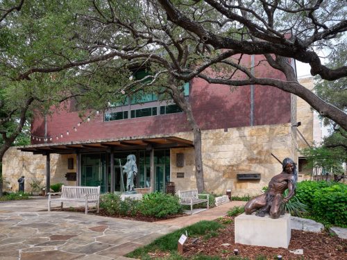 A building with natural stone walls and a large, sloping red roof, surrounded by mature trees. In front of the building, there is a paved area with benches and two bronze statues: one of a standing figure and another in a dynamic pose, possibly engaged in an activity like dancing or sports.